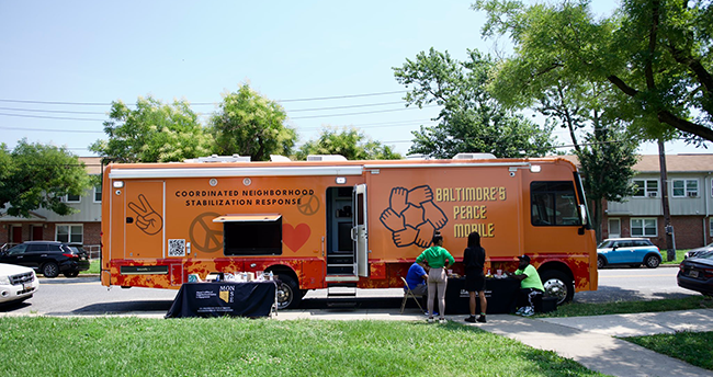 Photo of the "peace mobile" - an orange RV with text "coordinated neighborhood stabilization response" and "Baltimore Peace Mobile" on the side with a logo of six hands in a circle holding each other.